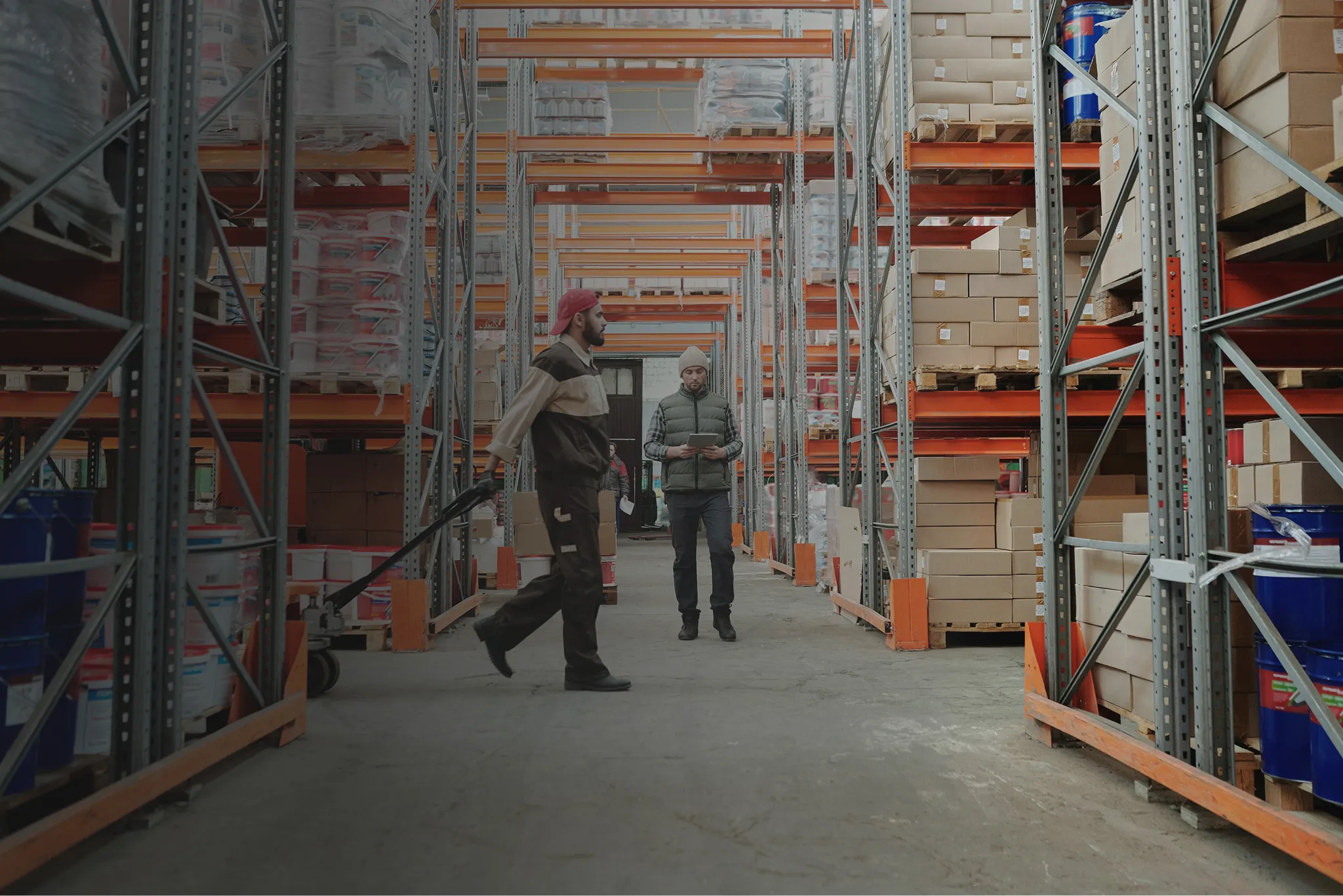 Two warehouse workers in aisles with metal shelves stacked with boxes and containers, one using a pallet jack and the other checking a tablet.