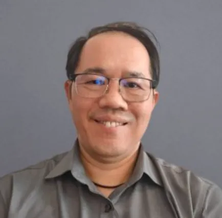 Smiling middle-aged man with glasses wearing a gray collared shirt against a plain gray background.