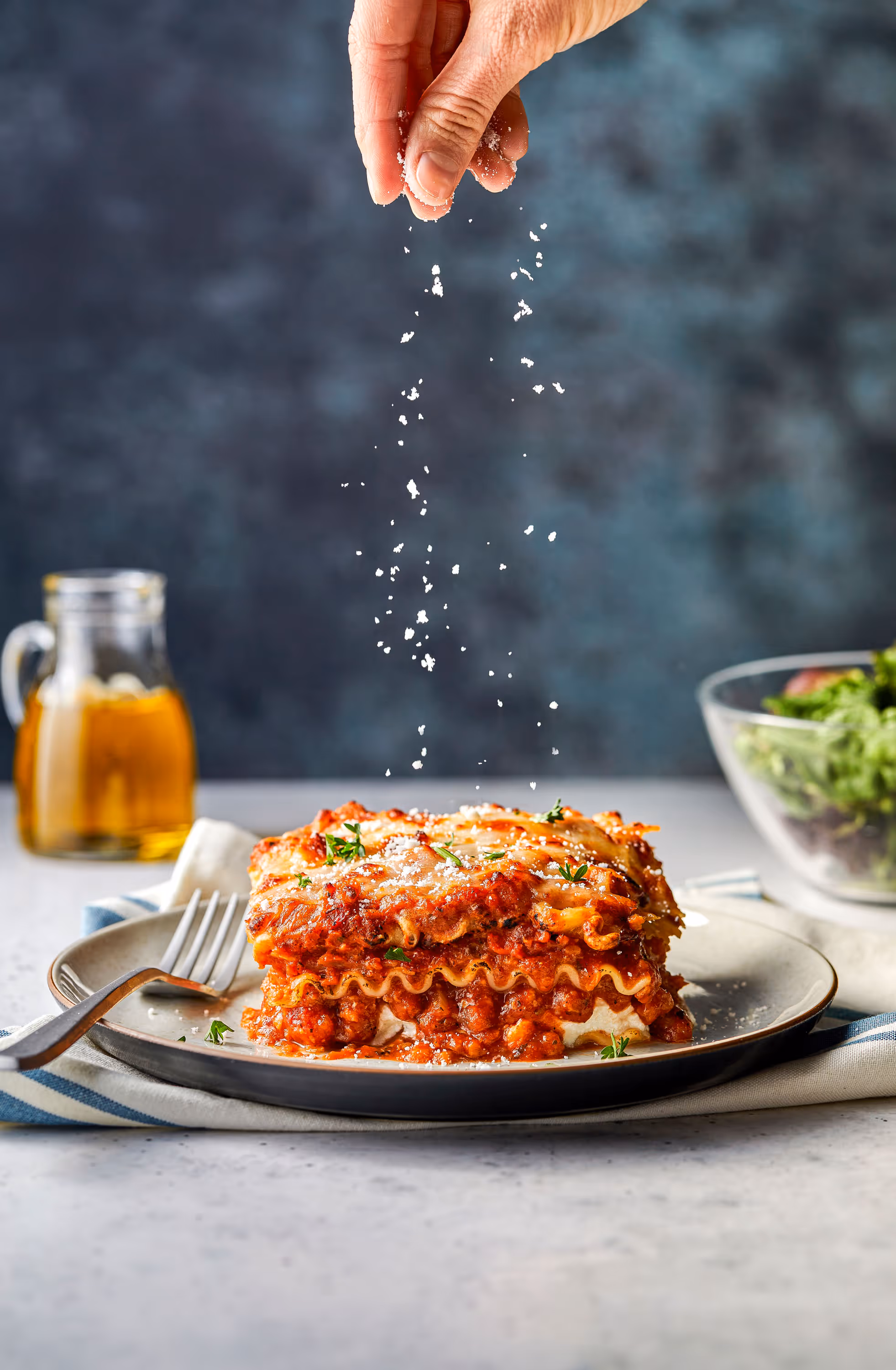 A photograph of a hand sprinkling parmesan from high above a serving of lasagna. Photography by Christi Chambers.