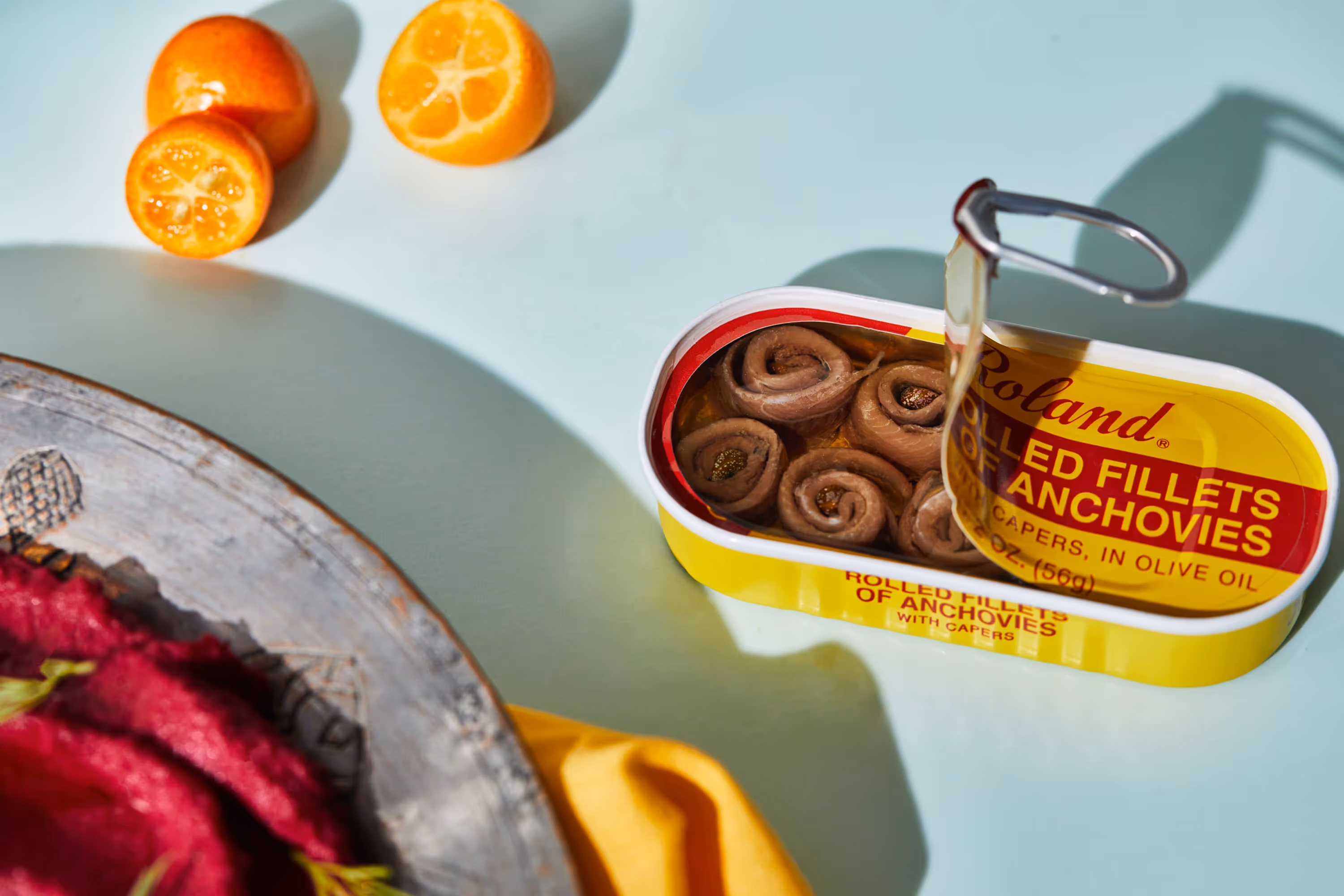 A tin of anchovies with the lid peeled halfway back, on a blue surface next to a plate of beet humus. Photography by Christi Chambers.
