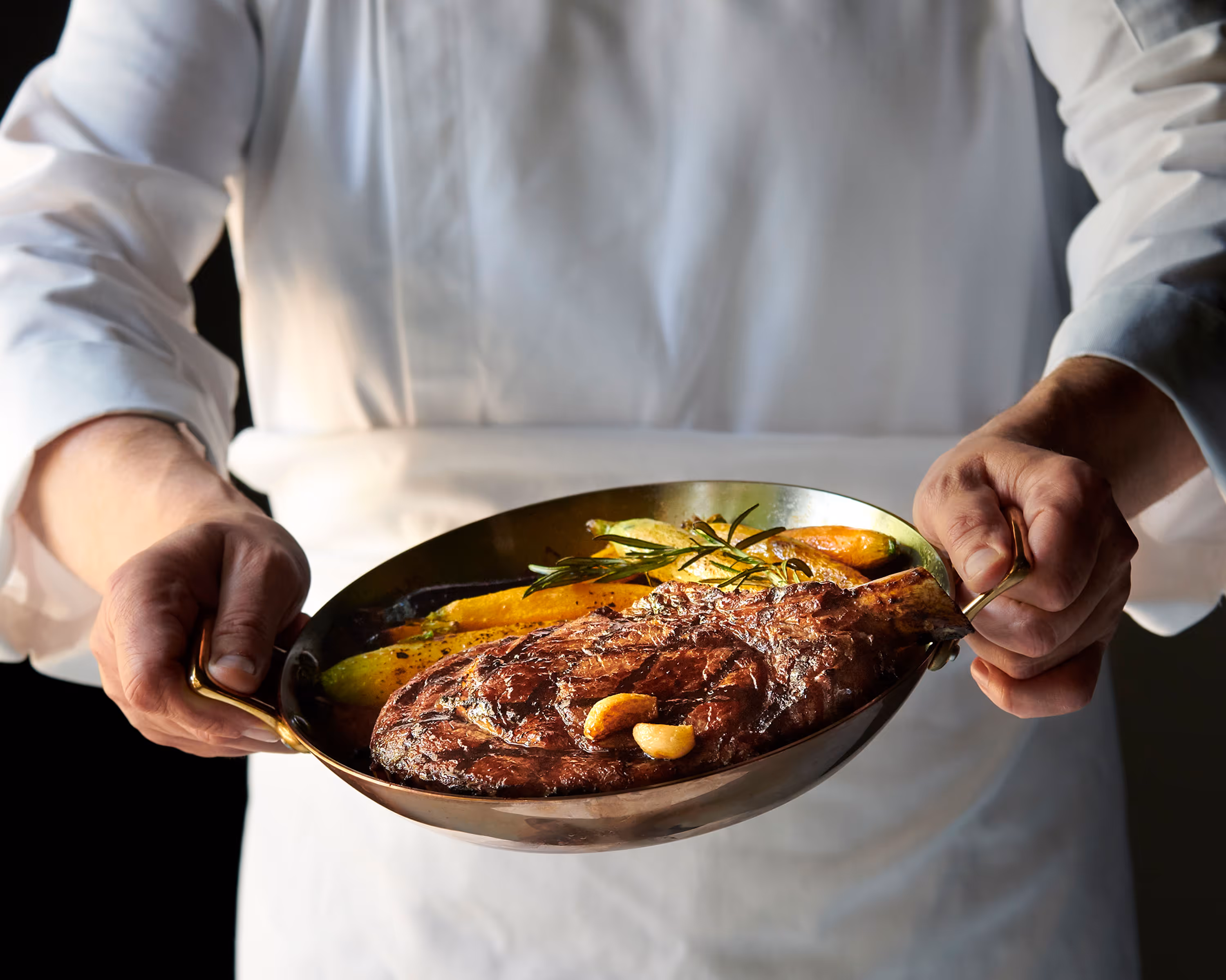 A moody photo of a chef holding a pan with a freshly cooked cut of meat, and vegetables. Photograph by Alex Crawford.