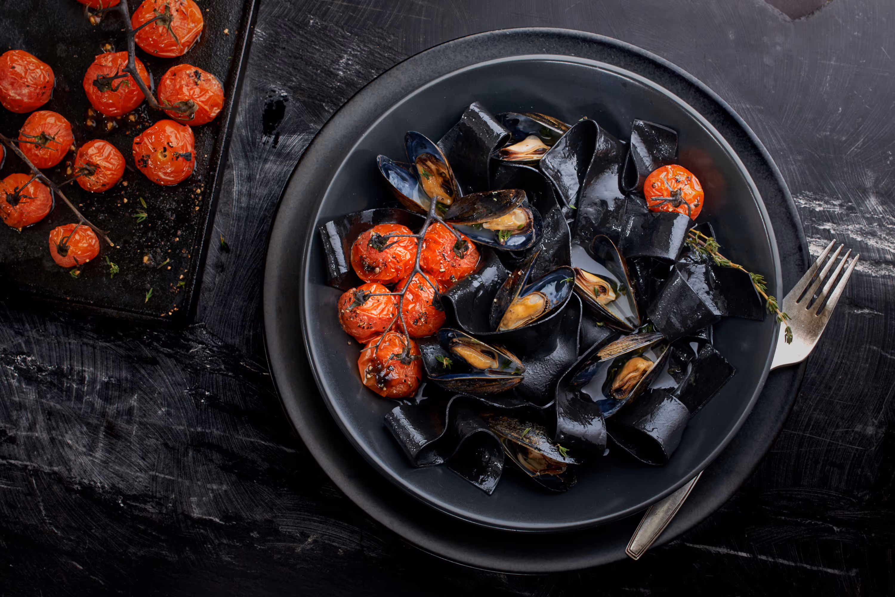 An overhead of a jet black squid ink pasta with sun-dried tomatoes. On a black plate with a black charger, on a dark surface. Photo by Alex Crawford.