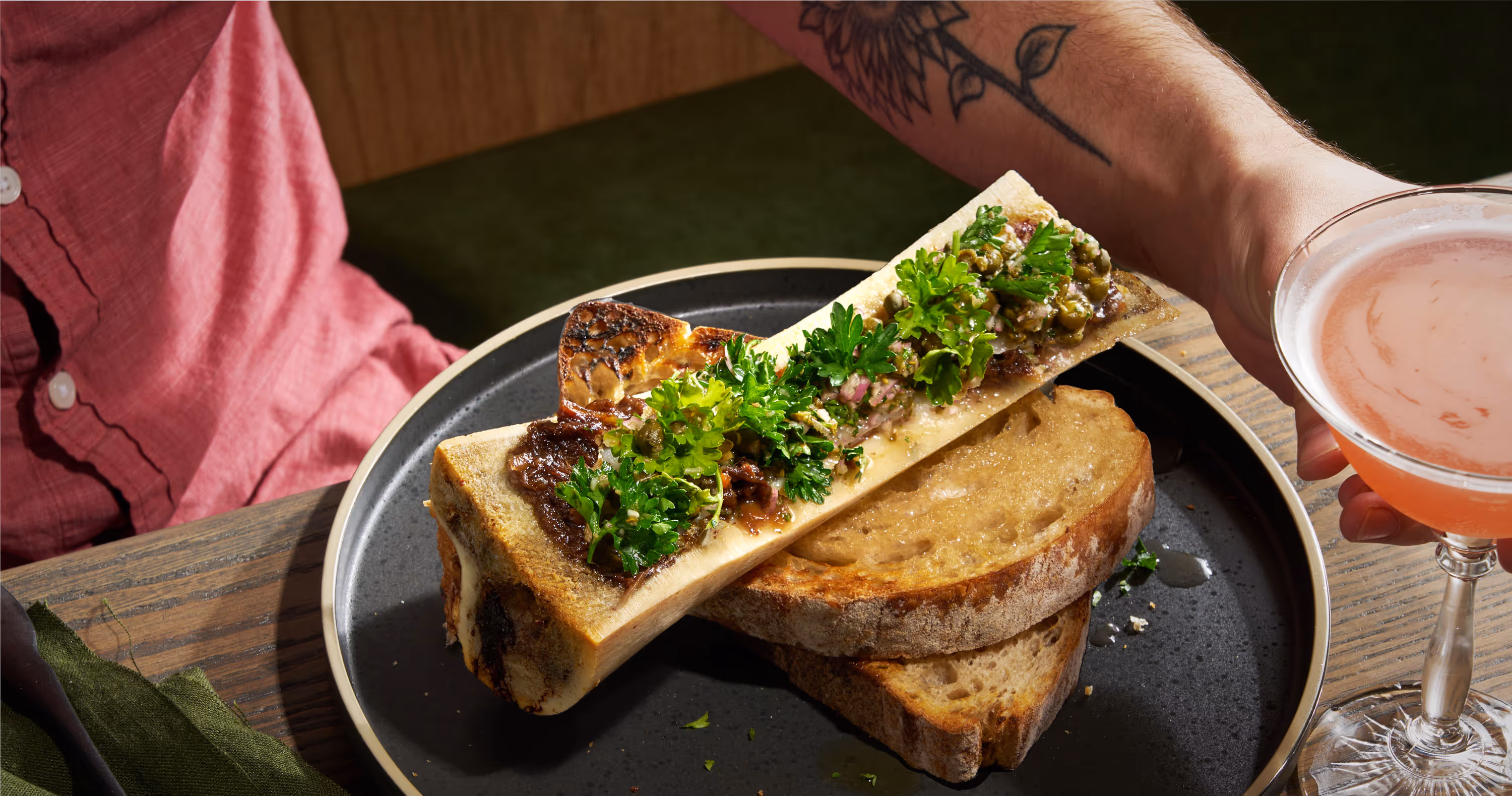 A photograph of a bone marrow salad, served in half of a beef bone, with texas toast, and a cocktail. Photograph by Amy Stallard.