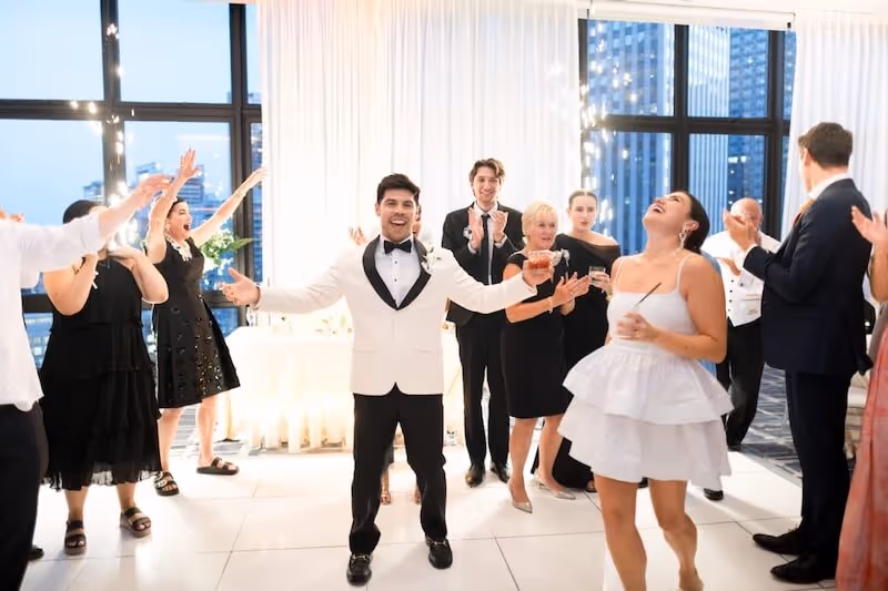 A happy groom in a white tuxedo in a white room with his bride and guests