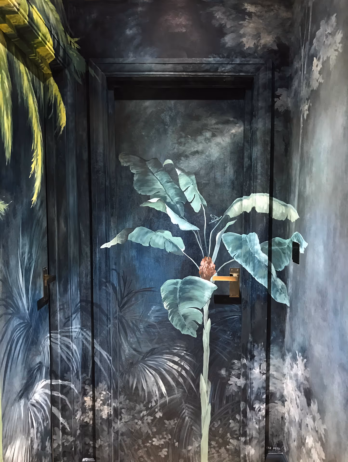 Floral and botanical mural by a London mural painter featuring a bright young palm against a dark tropical forest background, seamlessly blending wall and door into one elegant artwork.