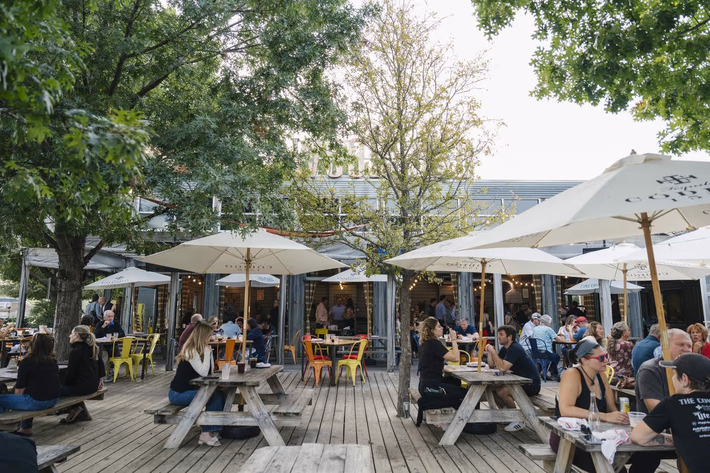 Crowded Woodshed Smokehouse patio with picnic tables, umbrellas, and dining under a tree canopy in daylight