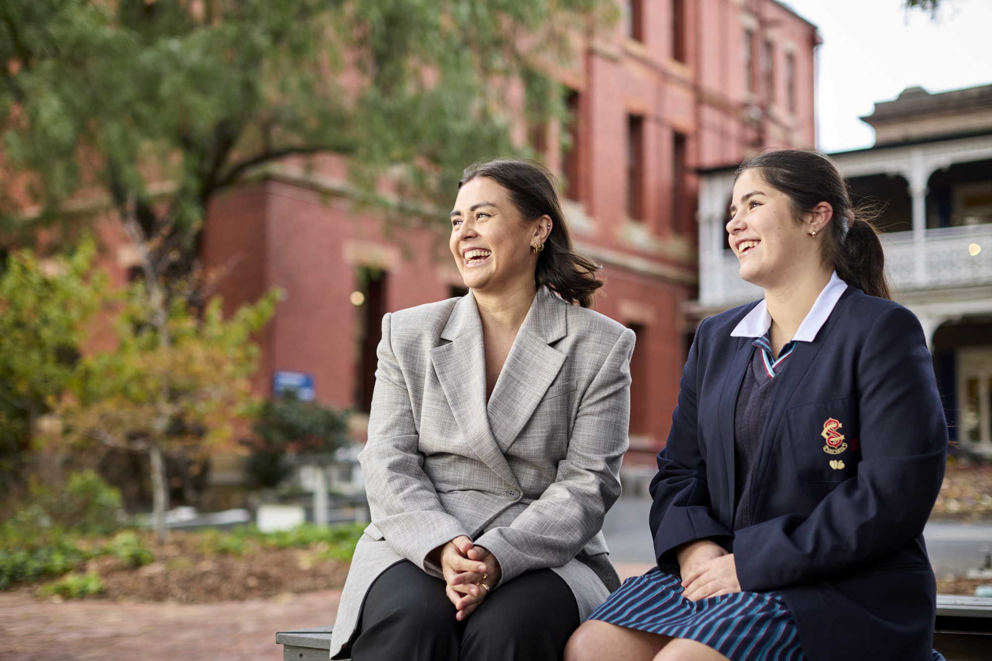 Two women sitting and smiling outside a red brick building with greenery around.