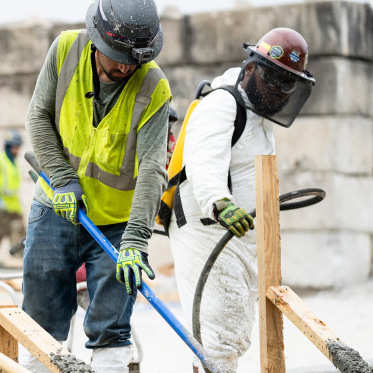 Construction workers pouring and leveling concrete at a job site, wearing safety gear including helmets, gloves, and protective clothing.
