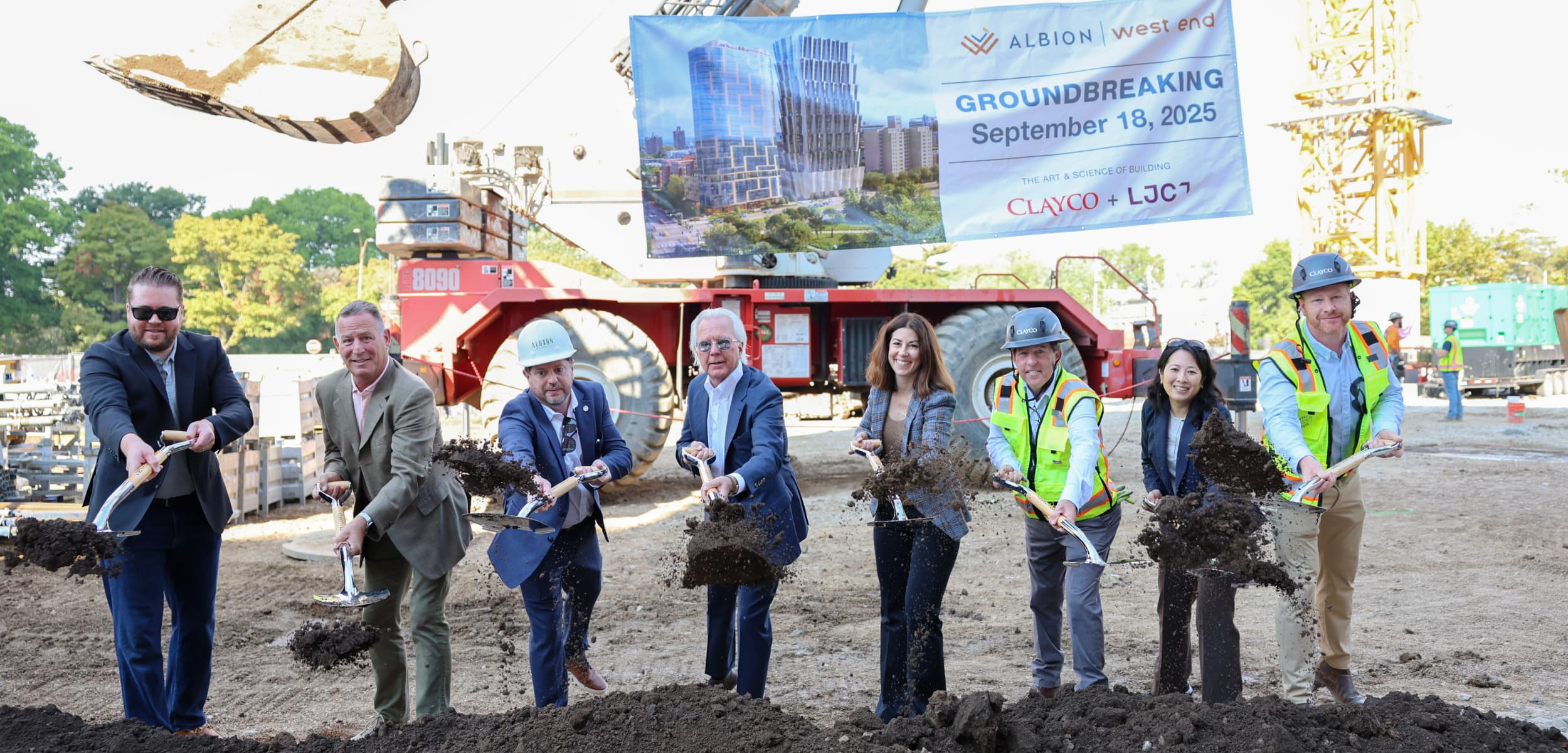 A group of eight people in business attire and construction gear participate in a groundbreaking ceremony at a construction site. They are holding shovels and tossing dirt, standing in front of construction equipment. Behind them hangs a large banner that reads: “ALBION | west end, GROUNDBREAKING, September 18, 2025. The Art & Science of Building. CLAYCO + LJC.” The banner also shows a rendering of a tall modern glass building.