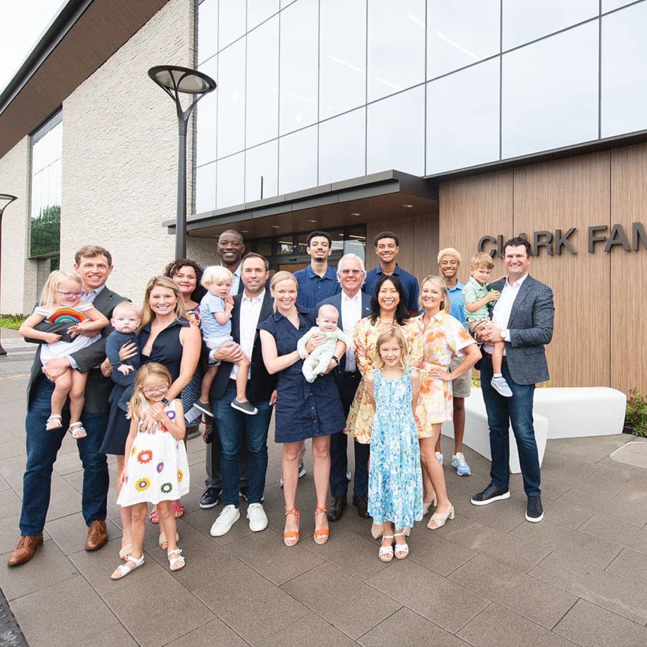 A large group of smiling family members and friends stand together outside the Clark Family Branch of the St. Louis County Library, posing for a photo in front of the modern glass-and-wood building.
