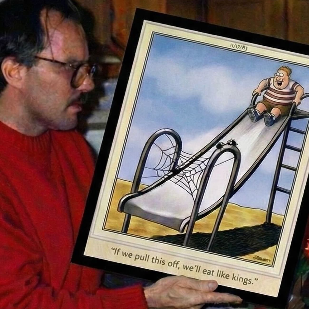 Bob Clark in a red sweater holds a framed Far Side cartoon showing a chubby child about to slide down a playground slide where two spiders have woven a web at the bottom; a caption reads, “If we pull this off, we’ll eat like kings.”