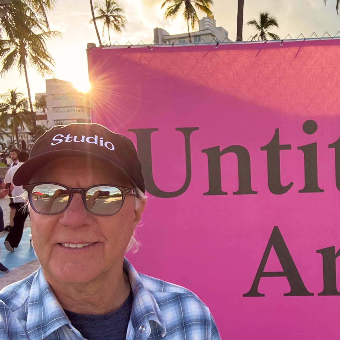 A man wearing sunglasses, a black "Studio" cap, and a plaid shirt takes a selfie in front of a bright pink sign that reads "Untitled Art," with palm trees and a sunset in the background.