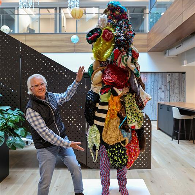 An older man with white hair, glasses, a plaid shirt, and a black vest poses enthusiastically beside a tall, colorful abstract sculpture made of layered textiles, beads, and various textured materials. He gestures toward the sculpture with a smile. The artwork stands on a white platform in a modern, open office space with wood floors, plants, a perforated metal staircase, and hanging decorative lights.