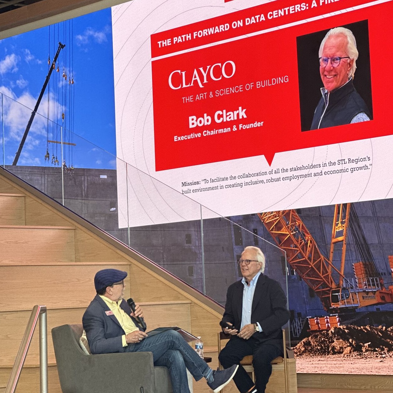 A conference stage scene where two men sit in armchairs during a fireside chat. A large screen behind them displays a red and white presentation slide reading “Construction Forum: The Path Forward on Data Centers,” featuring Clayco and Bob Clark, Executive Chairman & Founder, along with his photo. One man speaks into a microphone while the other listens, with construction imagery and a crane shown in the background.