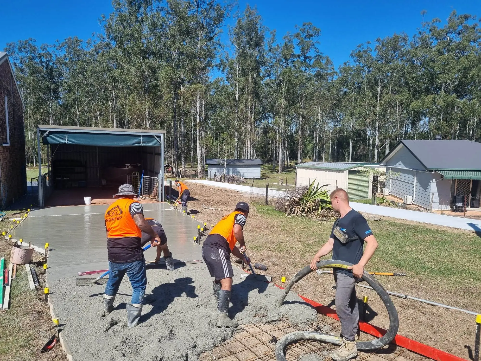 Four construction workers pouring and smoothing concrete driveway outside a house with trees in the background.