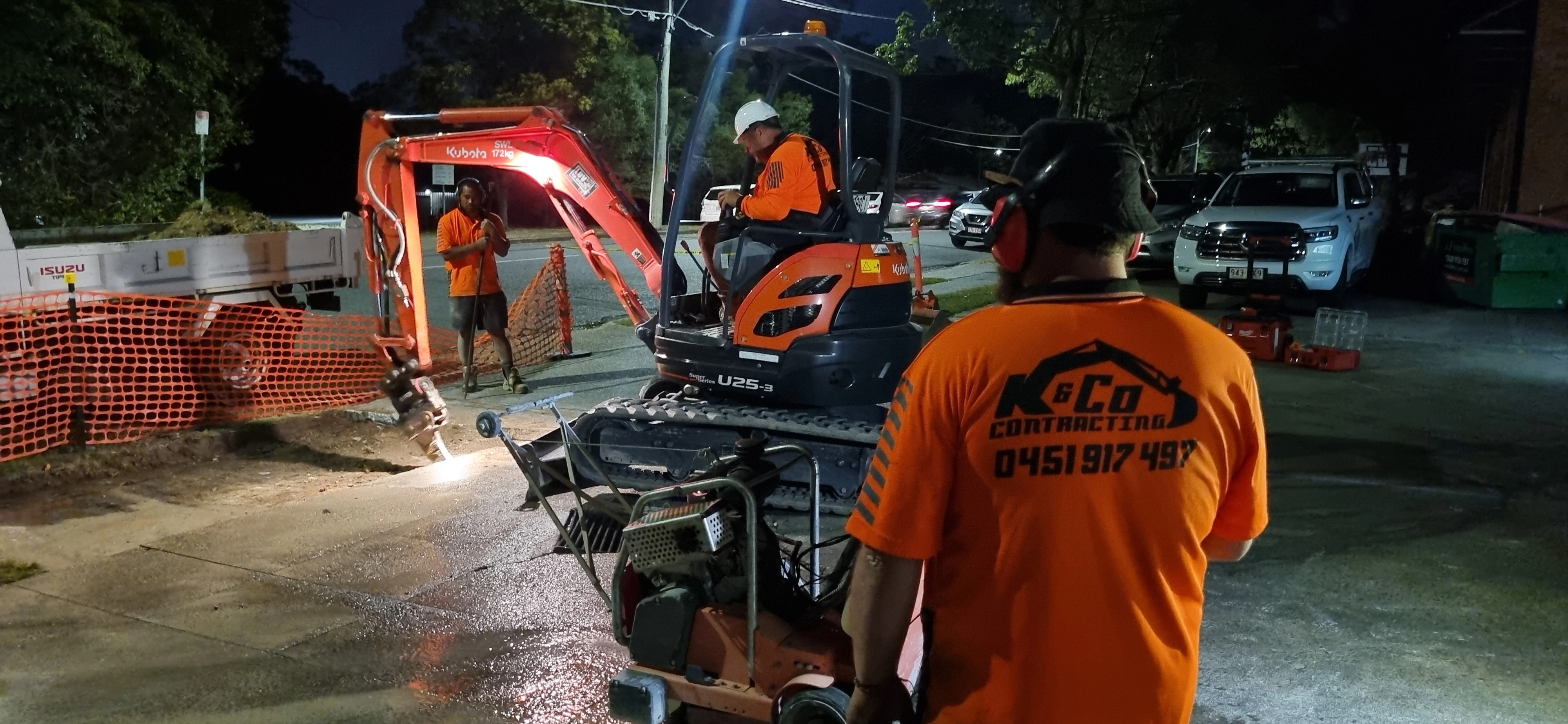 Construction workers in orange shirts operating an excavator and other equipment on a street at night.