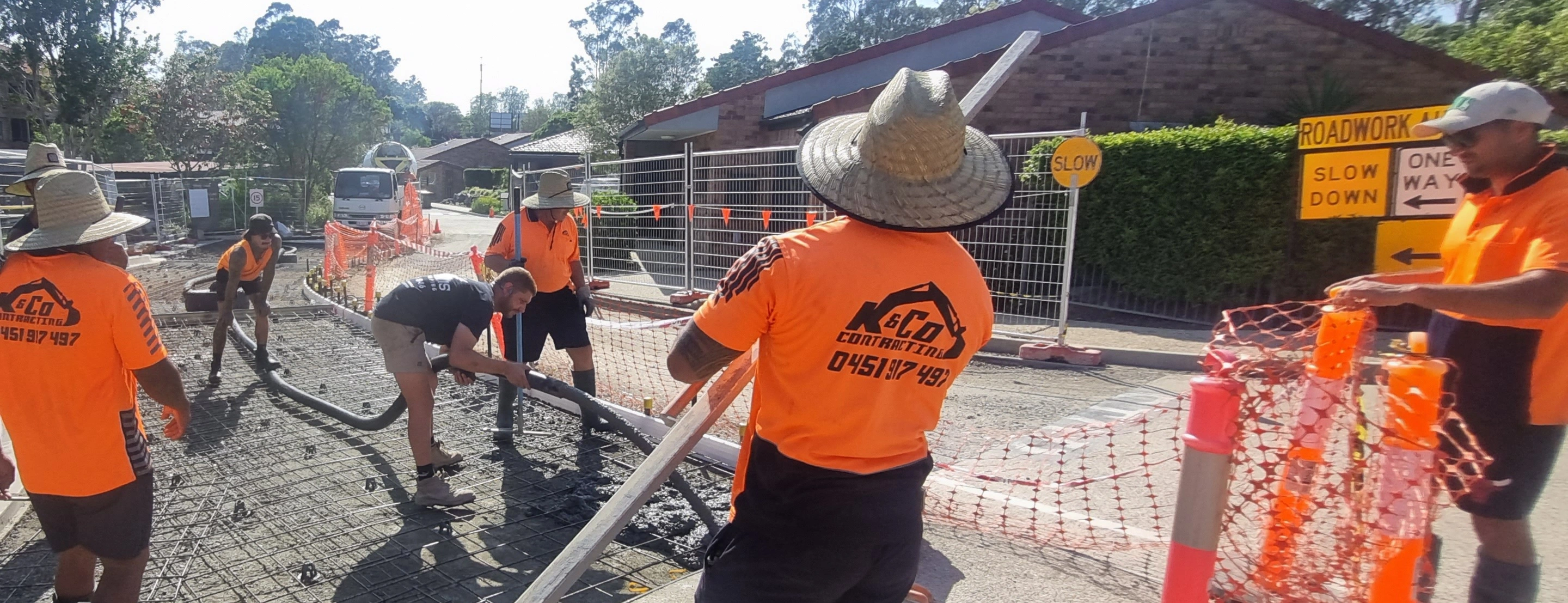 Construction workers in orange shirts and wide-brimmed hats pouring concrete on a road under sunny conditions with safety barriers and slow down signs.