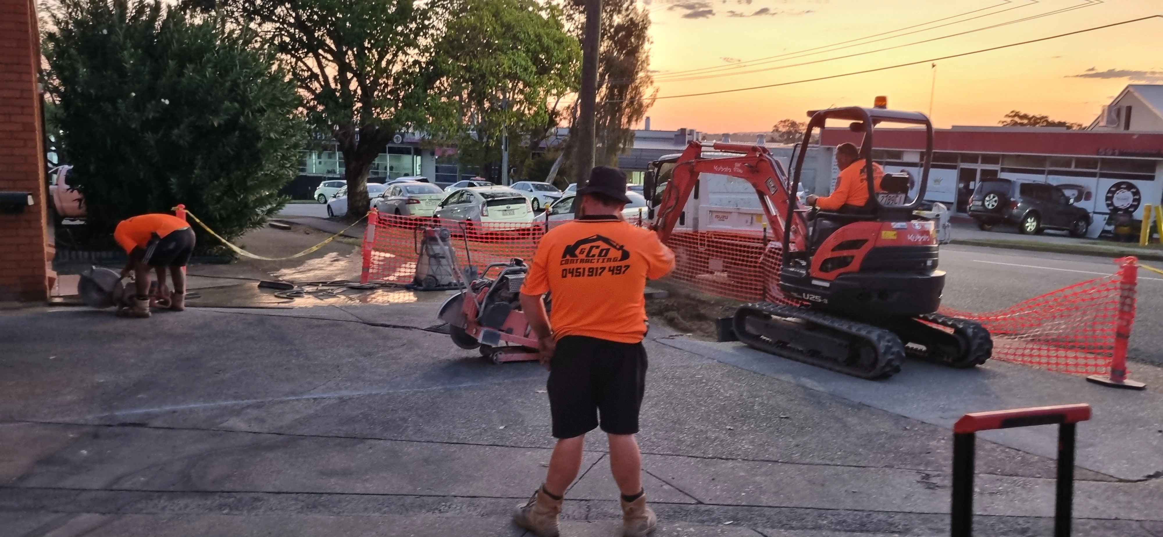 Three workers in orange shirts operating construction equipment on a roadside at sunset.
