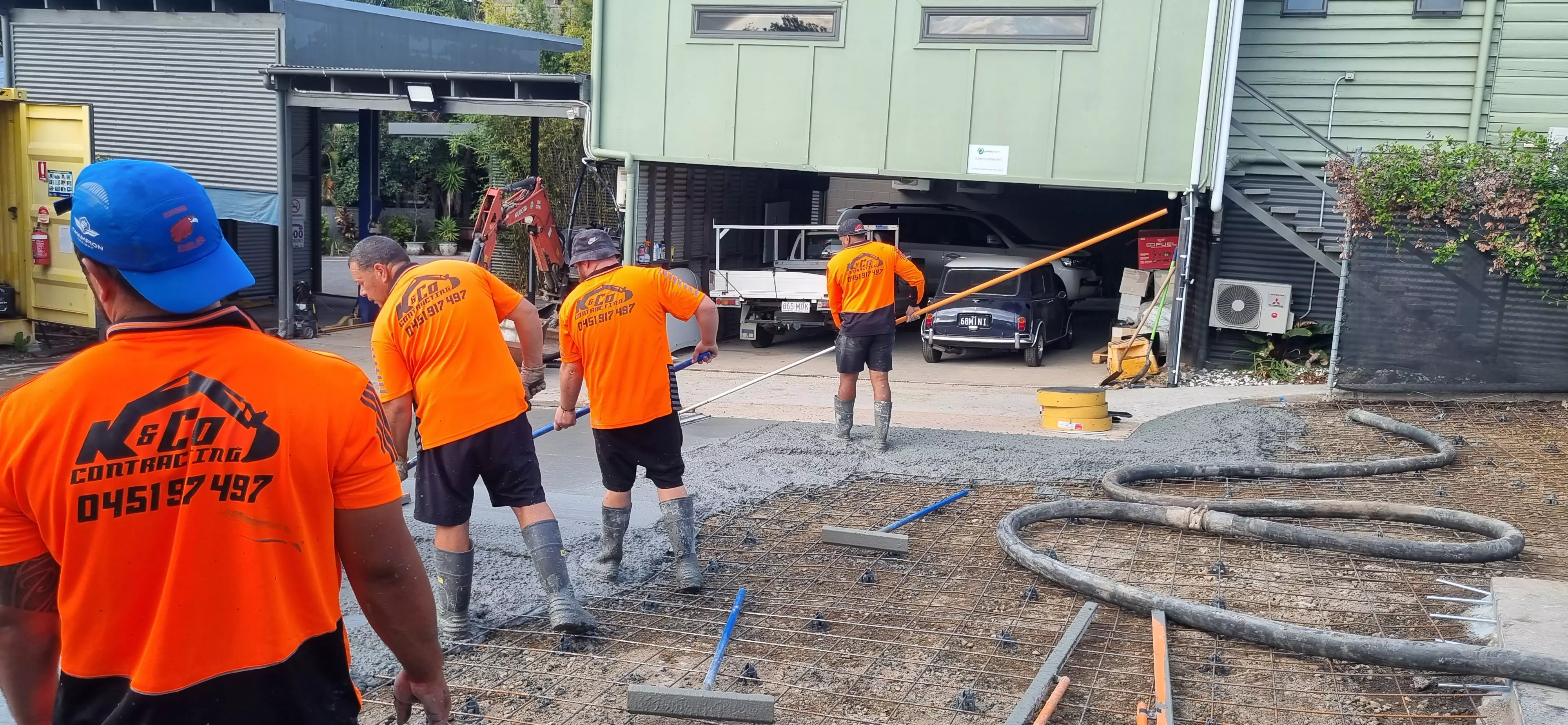 Four construction workers in orange shirts pouring and leveling concrete on a driveway near a garage with parked cars.