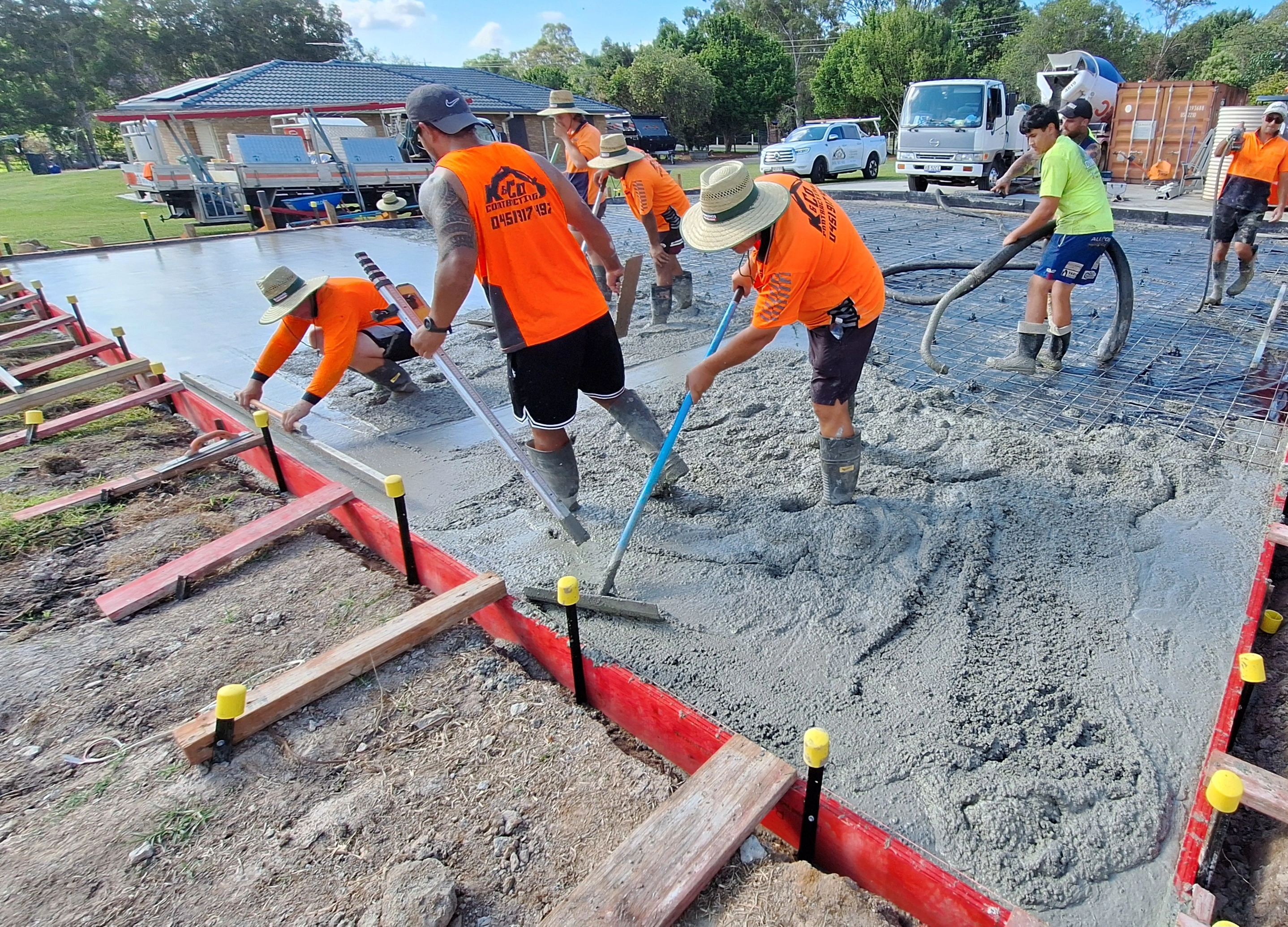 Construction workers wearing bright orange shirts and hats smoothing freshly poured concrete on a large outdoor slab.