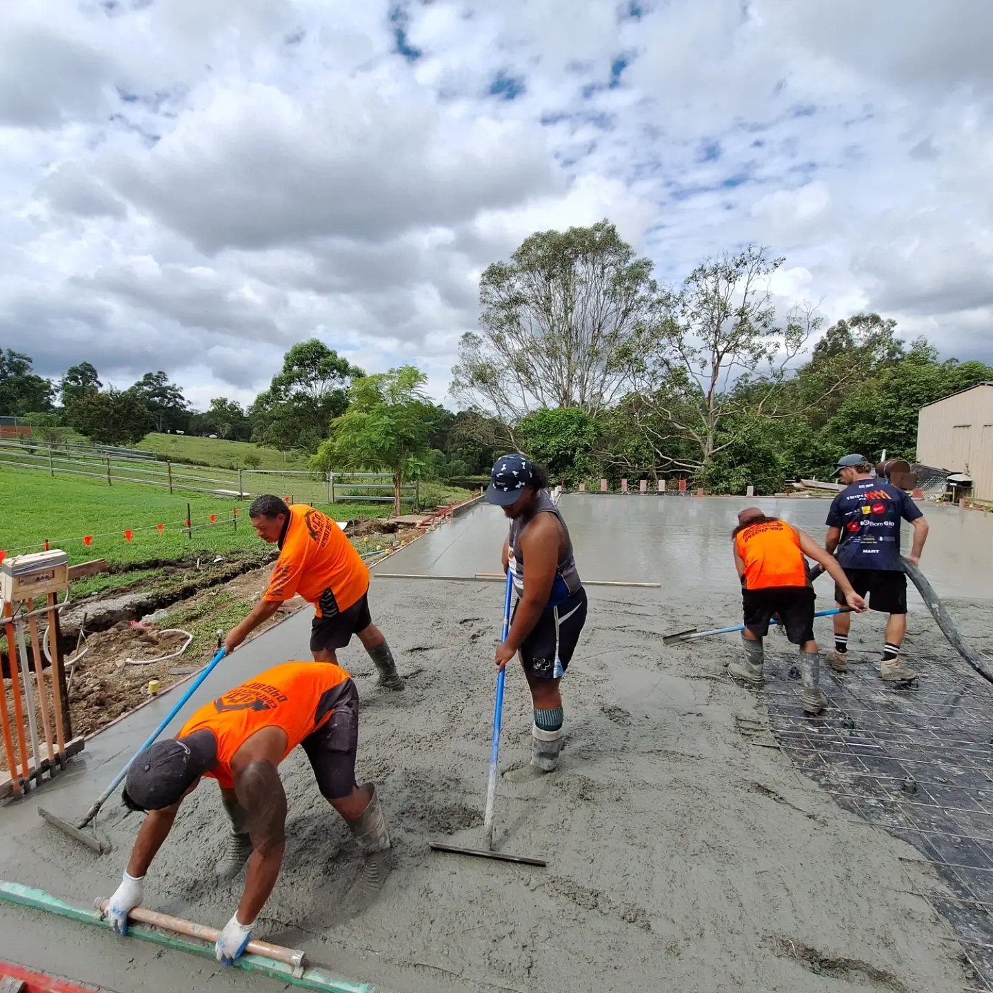 Five construction workers smoothing freshly poured concrete on an outdoor slab surrounded by green grass and trees under a partly cloudy sky.