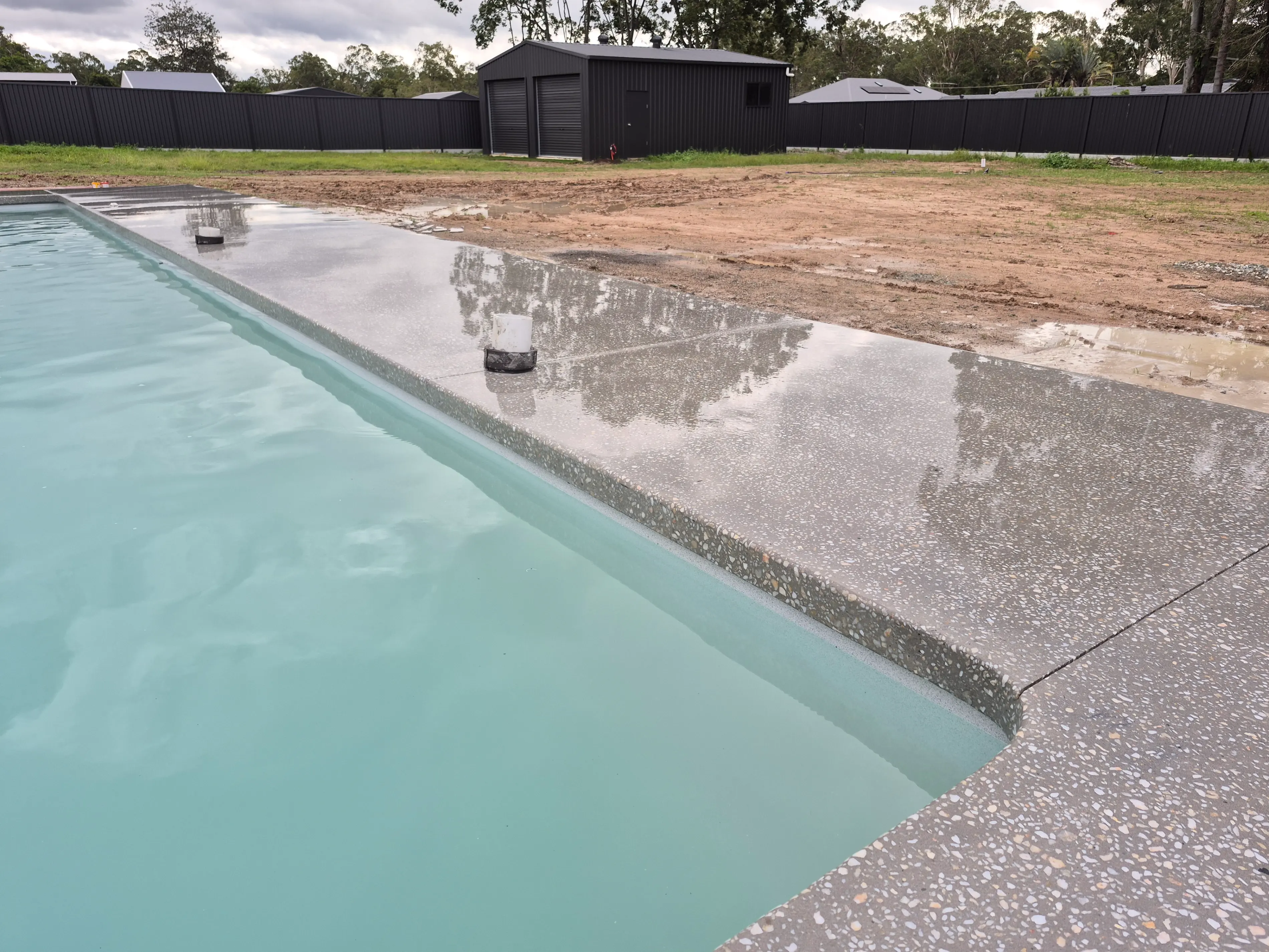 Edge of a light blue pool with wet exposed aggregate concrete decking, a black shed and fence in the background.