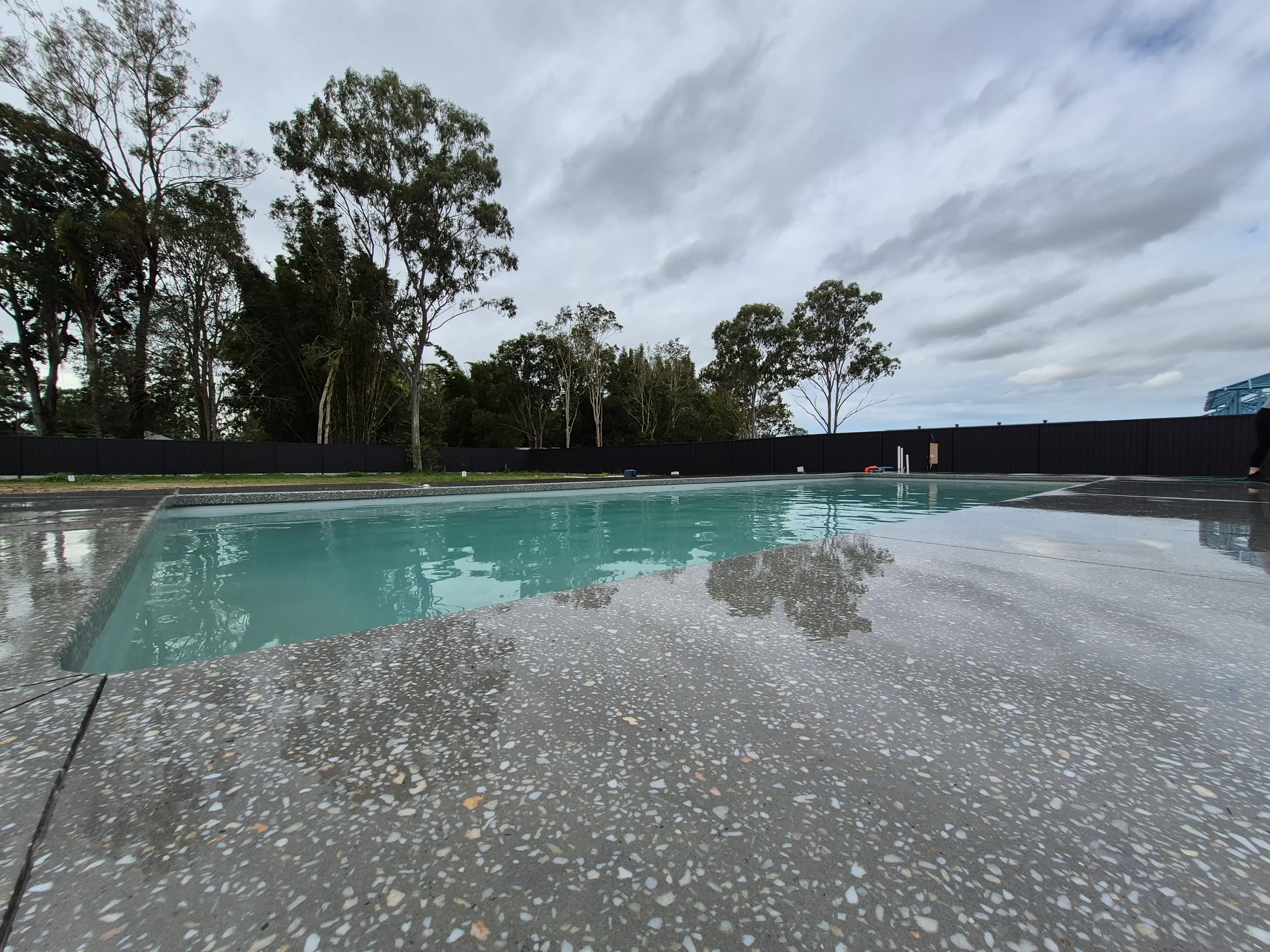 Outdoor rectangular swimming pool with clear blue water, surrounded by speckled concrete deck and trees under a cloudy sky.