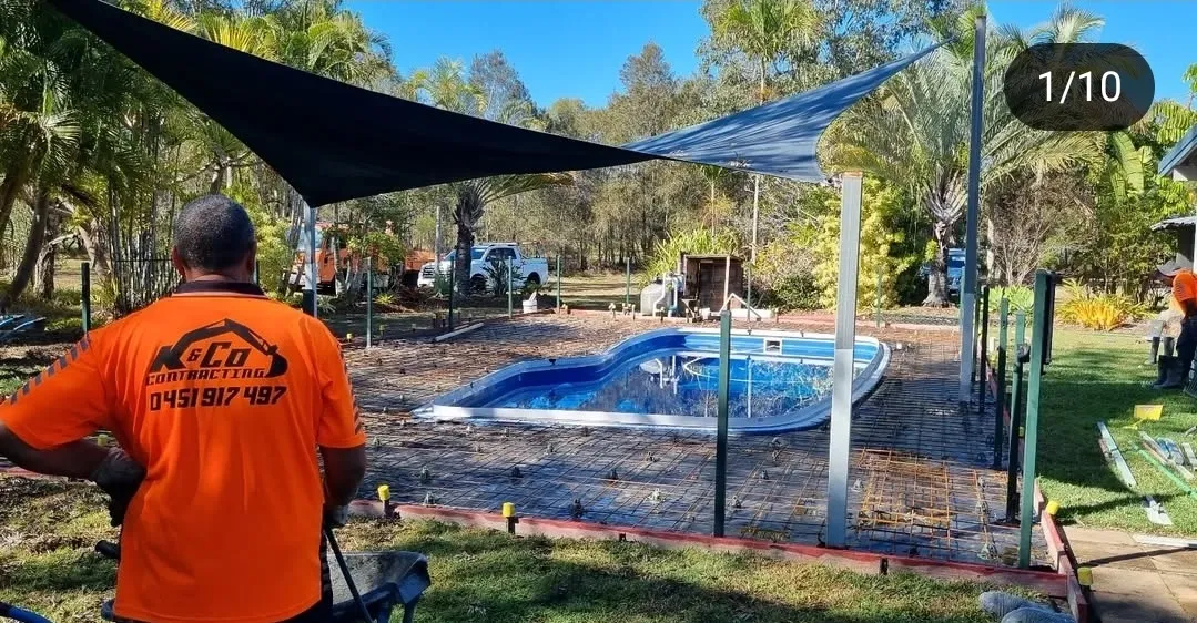 Construction workers wearing orange shirts setting up metal framework around a small backyard pool under black shade sails on a sunny day.