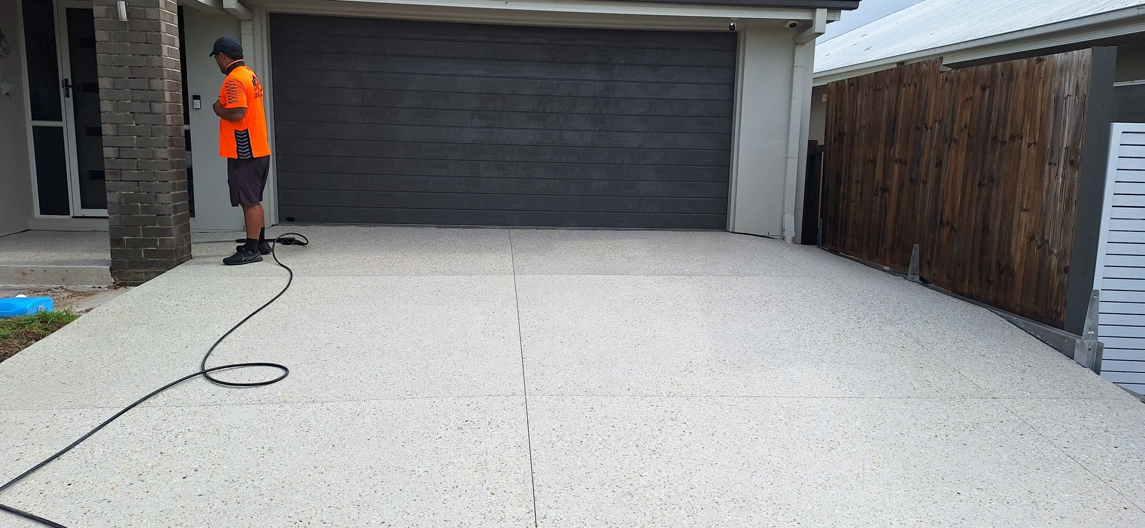 Man in an orange shirt standing at the entrance of a house with a dark gray garage door and light driveway.