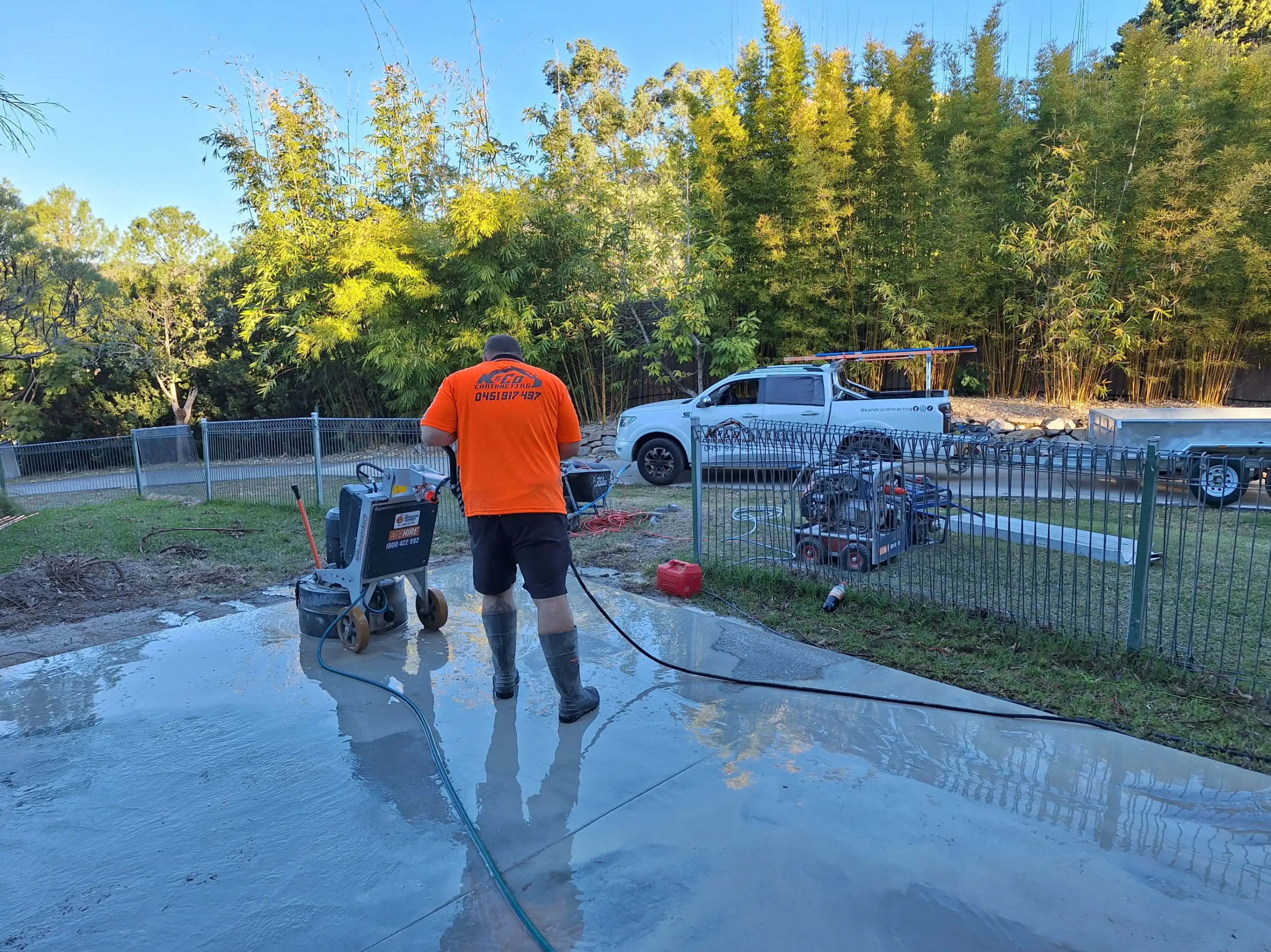 Worker in orange shirt and black shorts cleaning a wet concrete surface with a hose outdoors near a fence and parked white truck.