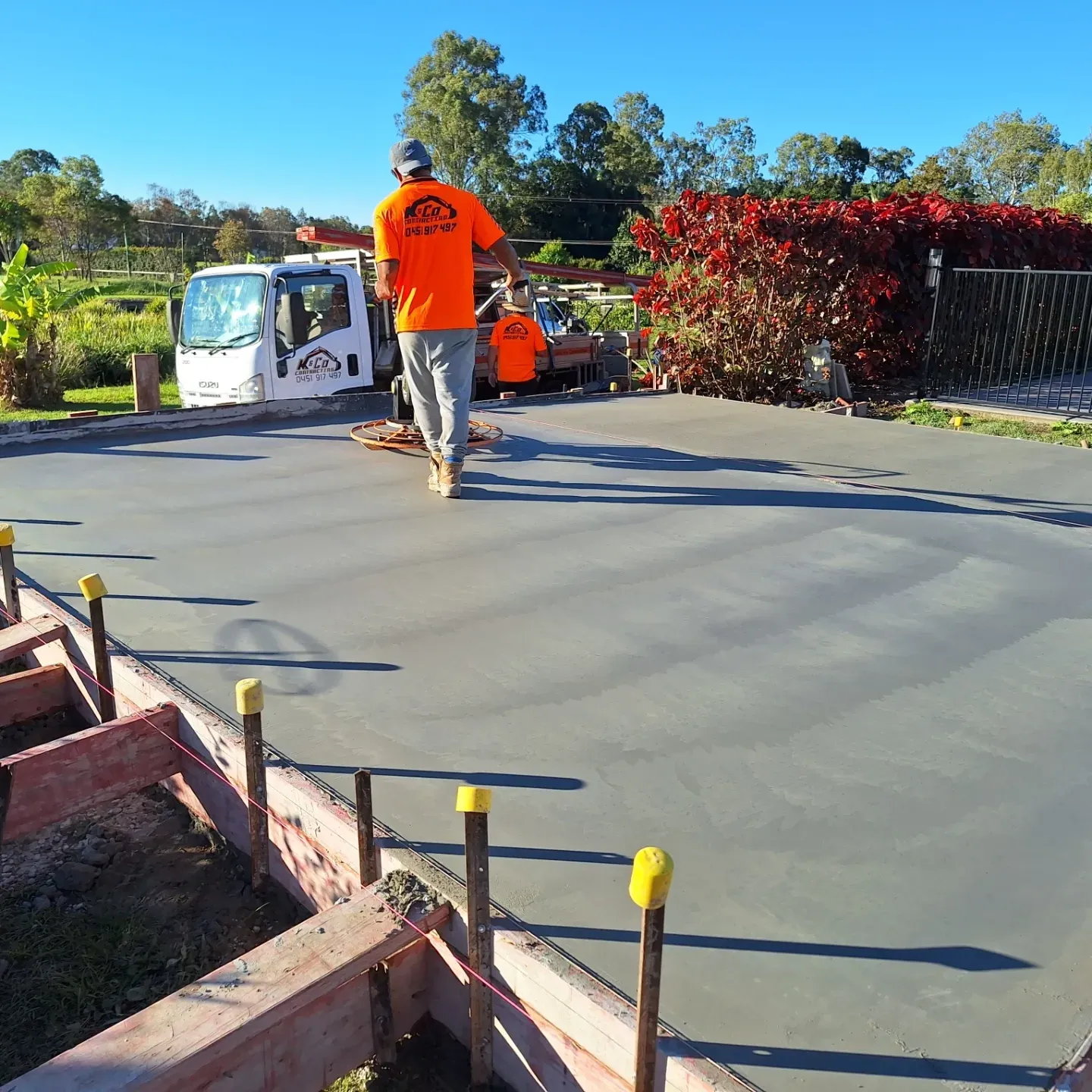 Construction workers smoothing a freshly poured concrete slab near a white truck on a sunny day.