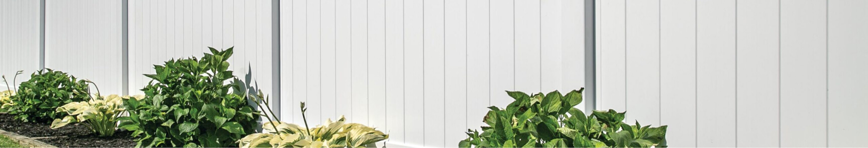 A white picket fence stands in front of a lush green garden under a sunny sky.