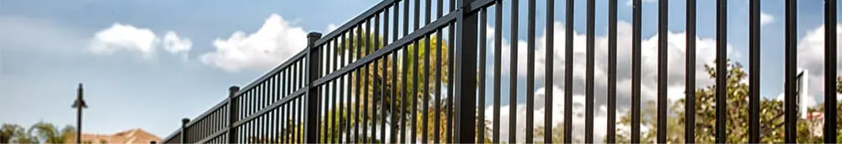 A black aluminum fence with evenly spaced vertical bars stands in front of a bright blue sky.