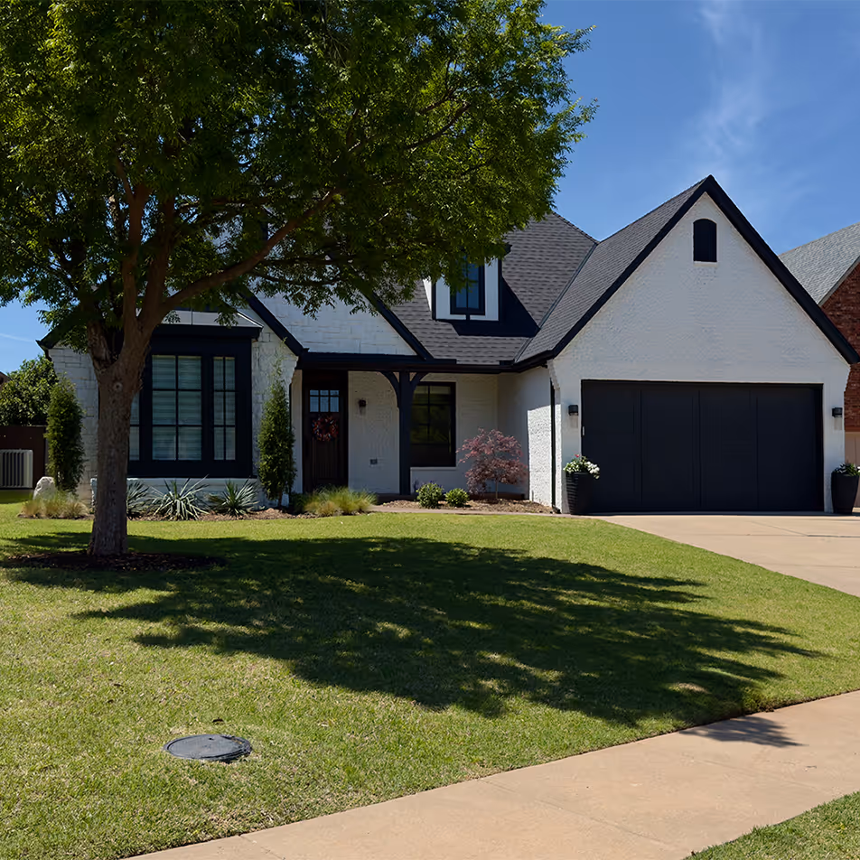 Modern white house with black trim, black garage door, tree casting shadow on green lawn, and a concrete driveway.