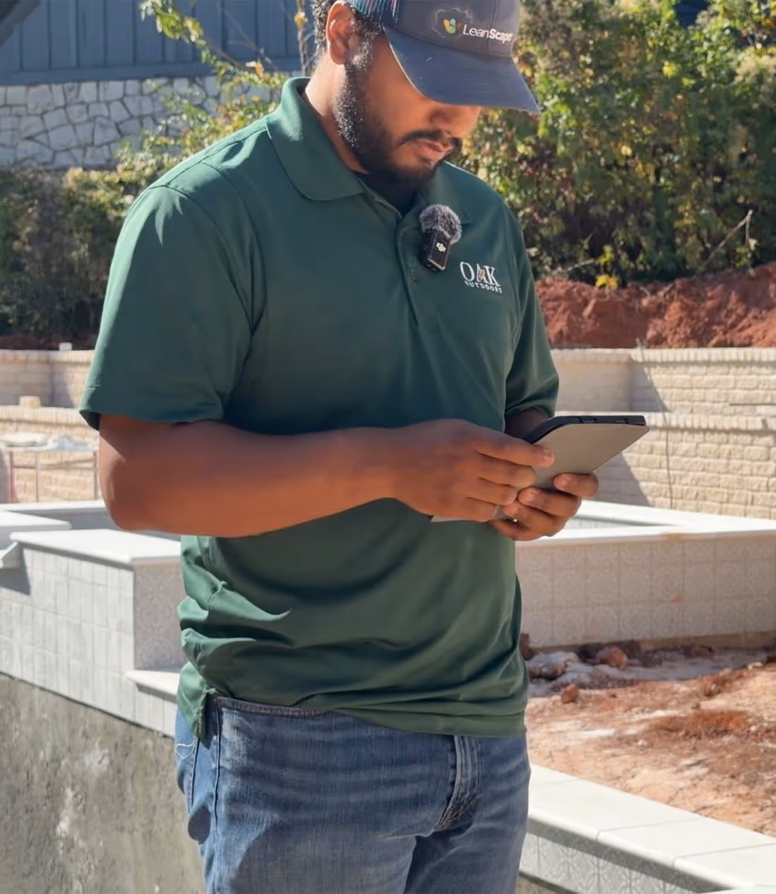 Man in green Oak Outdoors shirt and LeanScape cap using a tablet outdoors near construction materials.