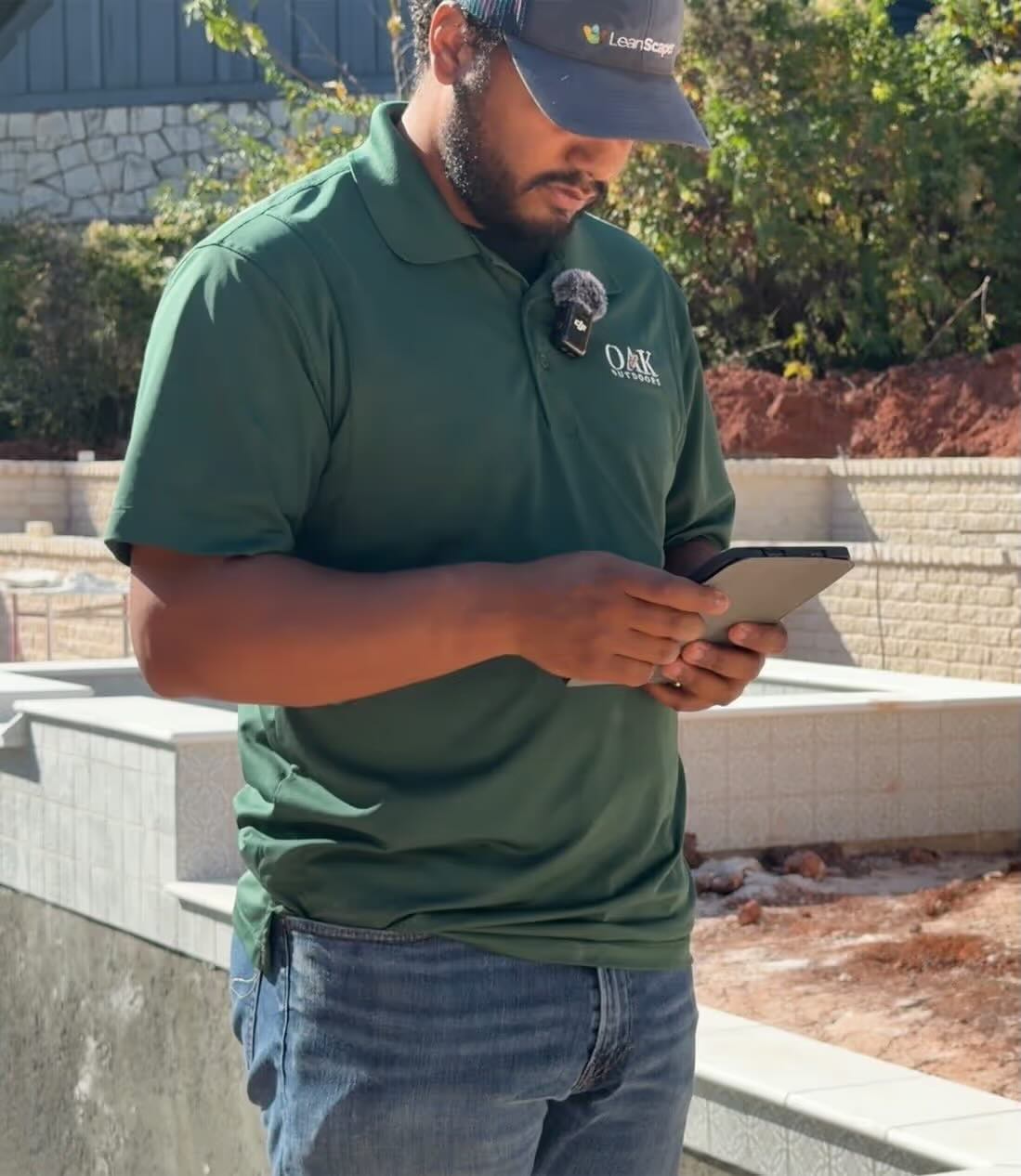 Man in green Oak Outdoors shirt and LeanScape cap using a tablet outdoors near construction materials.