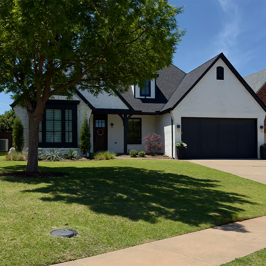 Modern white house with black trim, black garage door, tree casting shadow on green lawn, and a concrete driveway.