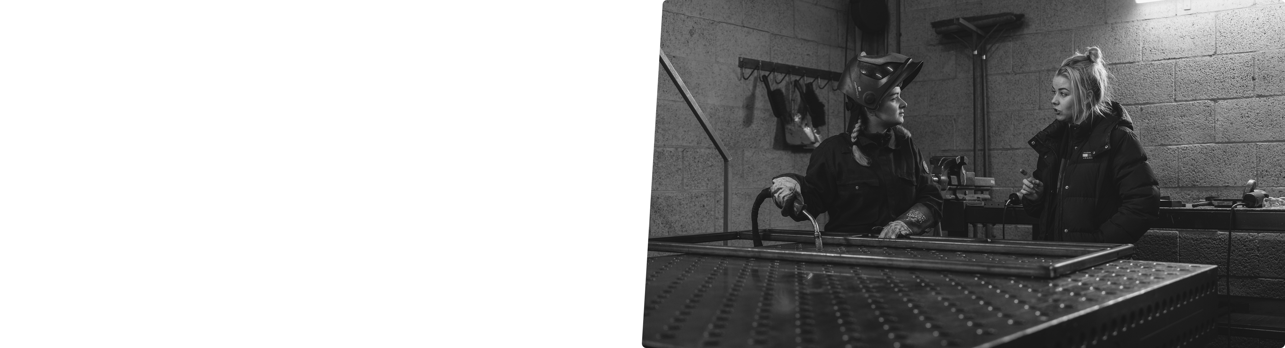 Black and white image of two women in industrial gear working and conversing in a workshop.