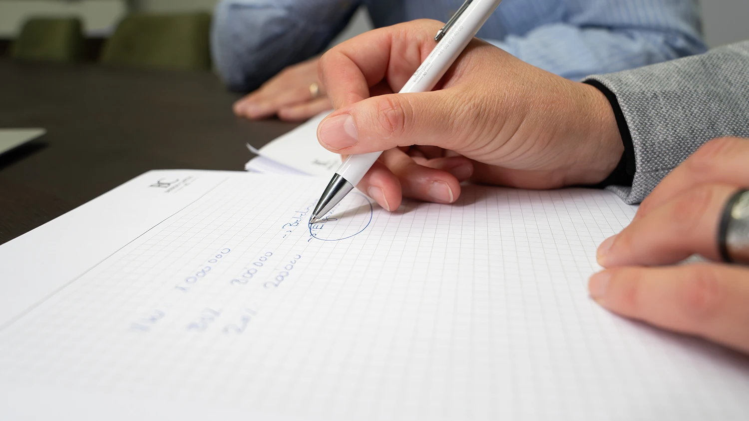 Nahaufnahme einer Hand mit einem Stift, die Zahlen und Notizen auf kariertem Papier schreibt.
