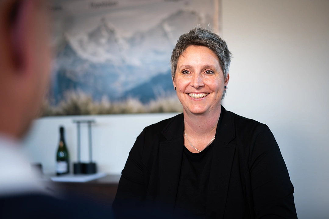 Lächelnde Frau mit kurzen grauen Haaren in einem schwarzen Blazer in einem Büro mit einem verschwommenen Bild von Bergen im Hintergrund.