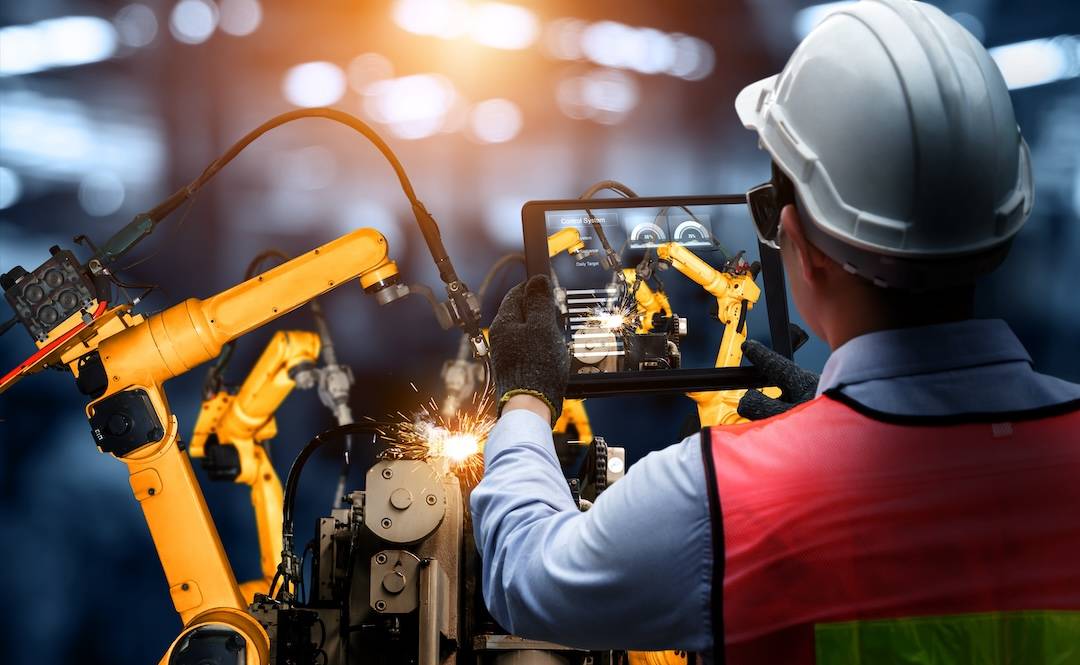 Man in safety helmet holding tablet displaying digital production software in front of robot arms in factory