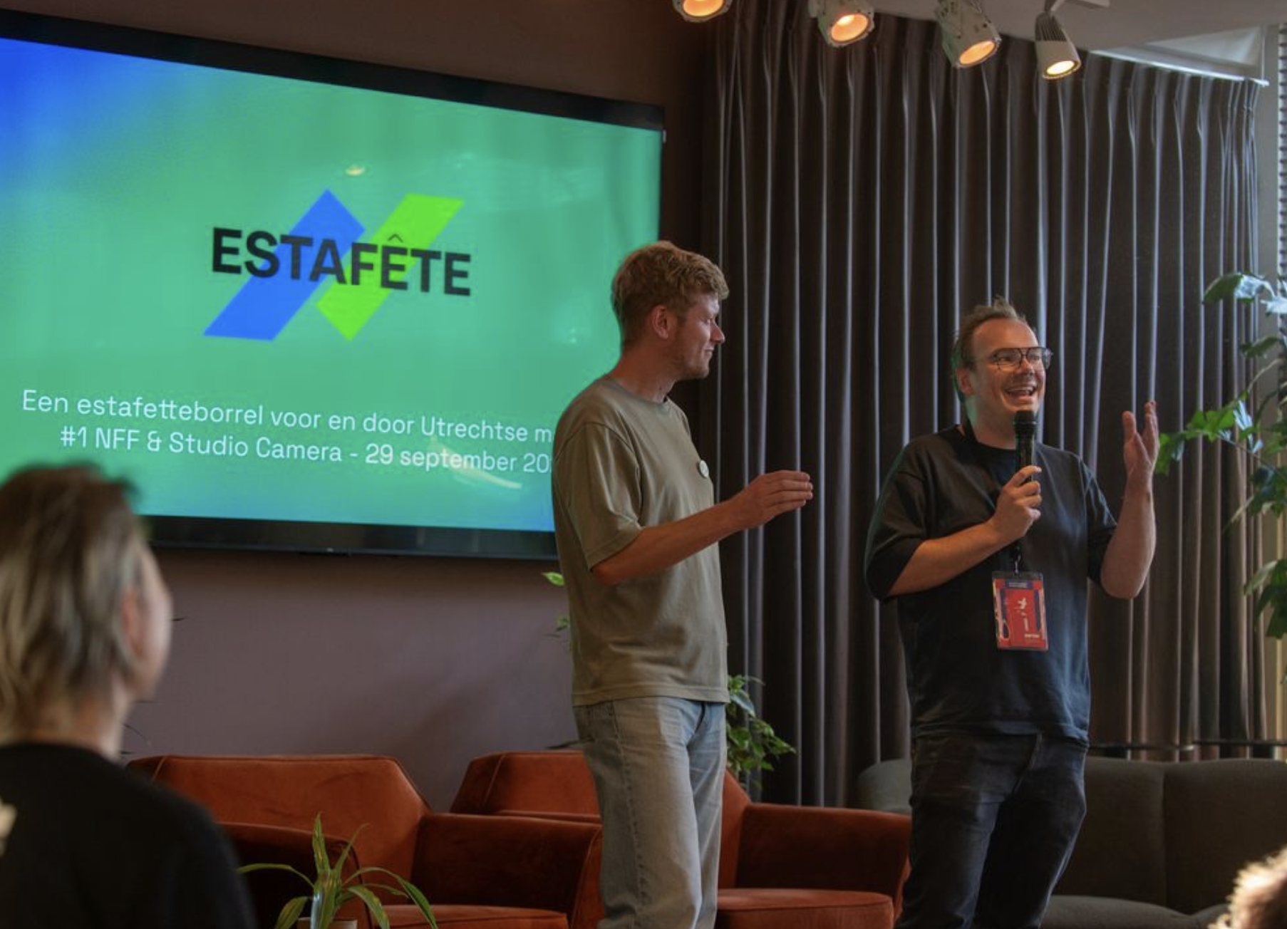 Twee mannen die presenteren voor een scherm en zaal met borrelende mensen