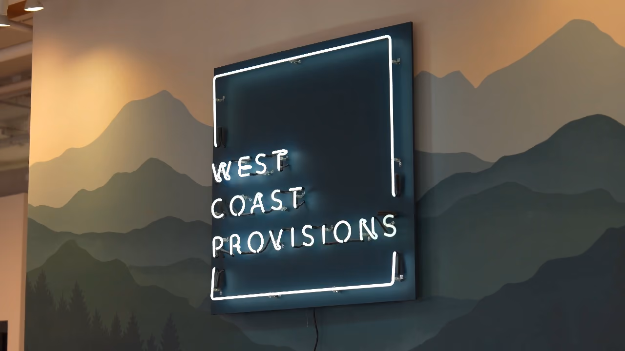 A neon sign on a wall with a painted mountain landscape in shades of green and blue, reading 'WEST COAST PROVISIONS' enclosed in a rectangular neon frame.