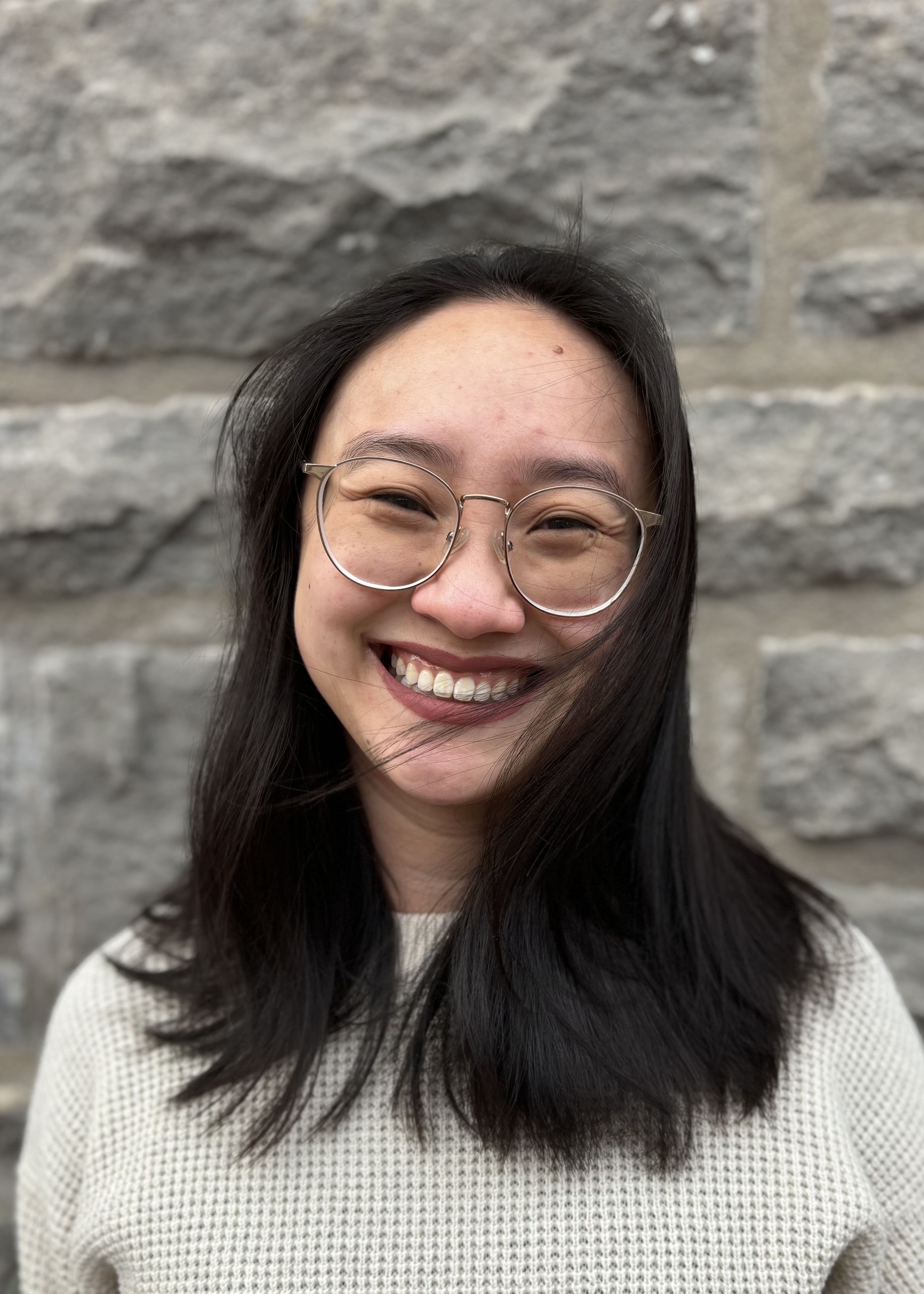 A portrait style photo of Emma Sue, the designer of this website. She has her dark hair down, about shoulder length. She is wearing a white knitted top and stands in front of a stone wall.