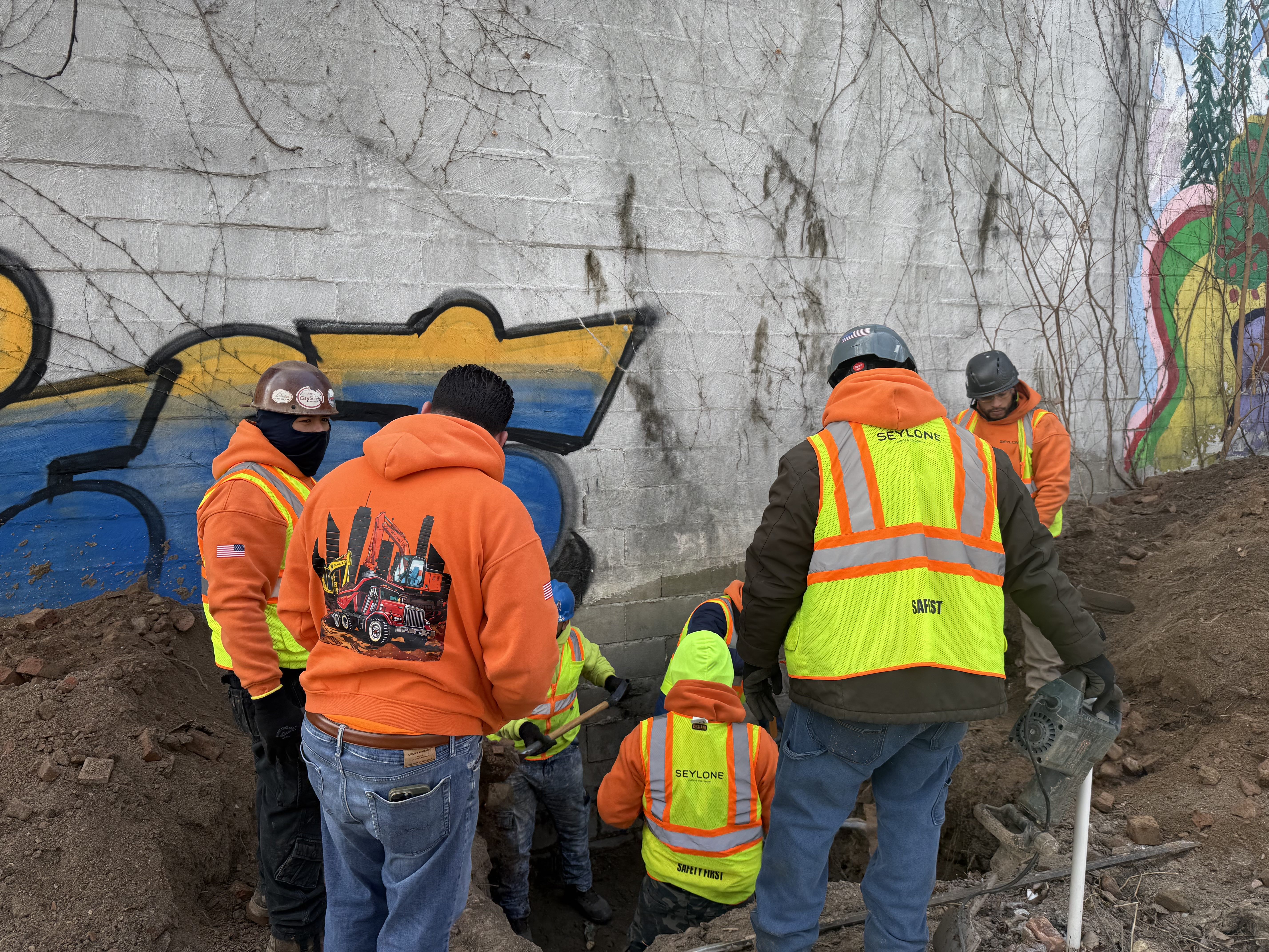Construction workers wearing orange hoodies and yellow safety vests work in a dug-out area beside a graffiti-covered concrete wall.