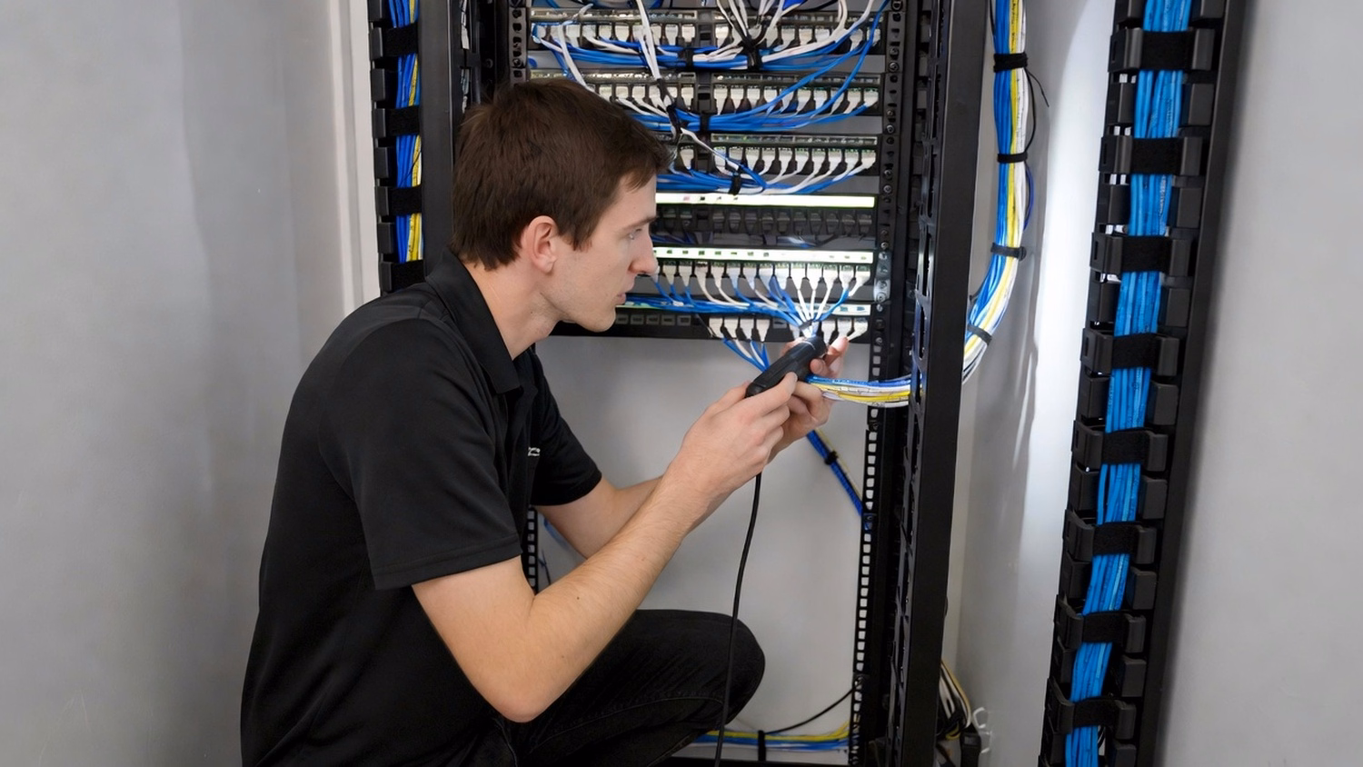 An engineer crouches in a network closet, tracing a cable run on a patch