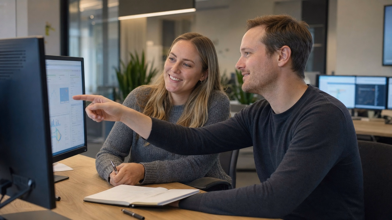 Two colleagues sit side-by-side at a shared table, reviewing information display