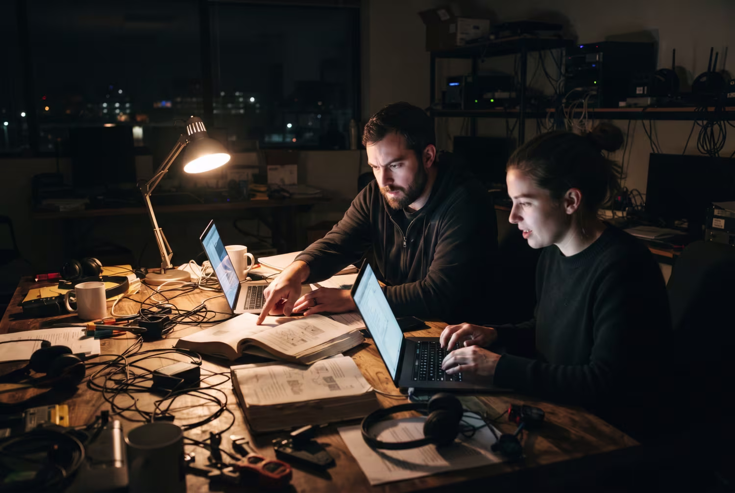 Two IT professionals collaborating at a cluttered desk in dimly lit office