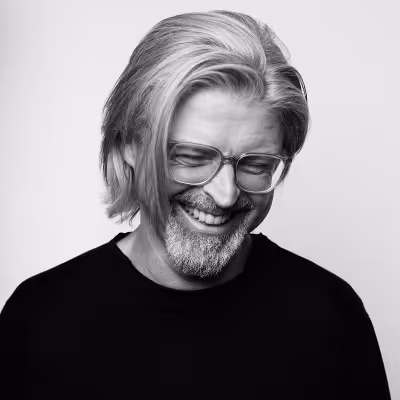 Smiling middle-aged man with gray hair, beard, and glasses wearing a black shirt against a plain background.