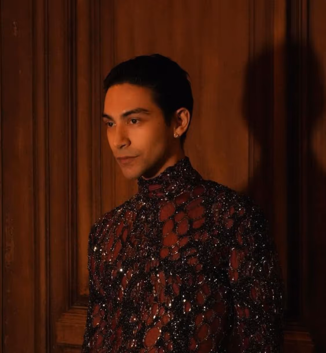 Man standing against wooden paneled wall wearing a dark red and black sequined high-neck shirt.